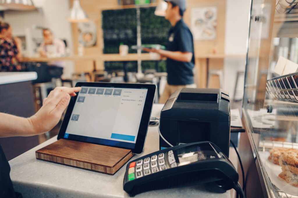 Small business owner working on a computer with a point of sale machine, blurred foot traffic in the background.