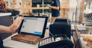 Small business owner working on a computer with a point of sale machine, blurred foot traffic in the background.