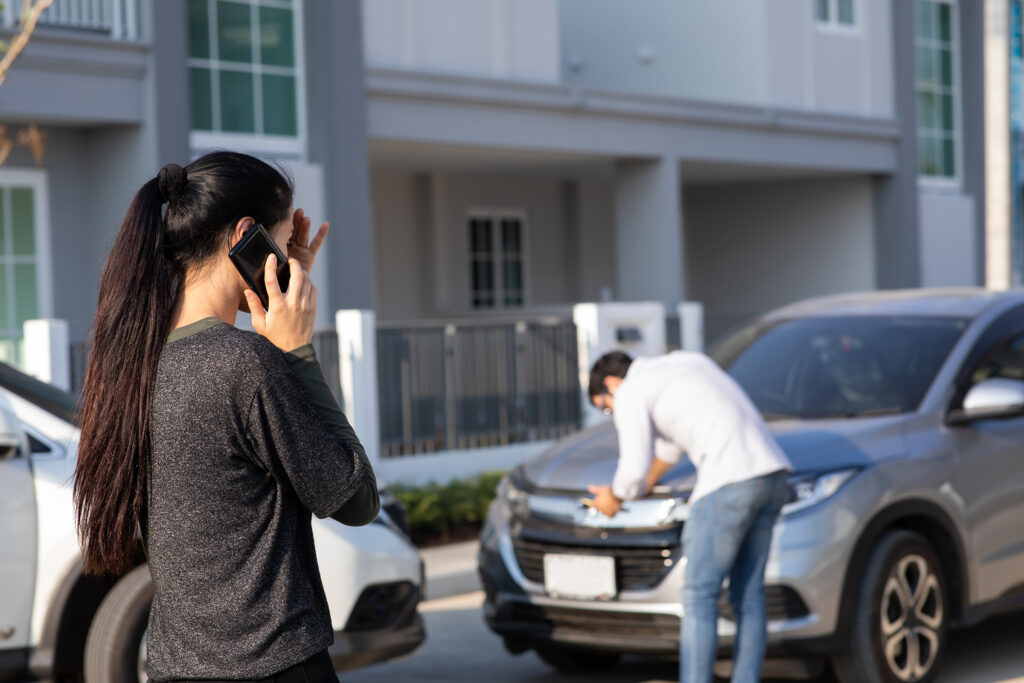 Woman making a phone call to her insurance agent after a car accident, while another driver inspects the roof of his vehicle in the background.