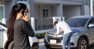 Woman making a phone call to her insurance agent after a car accident, while another driver inspects the roof of his vehicle in the background.