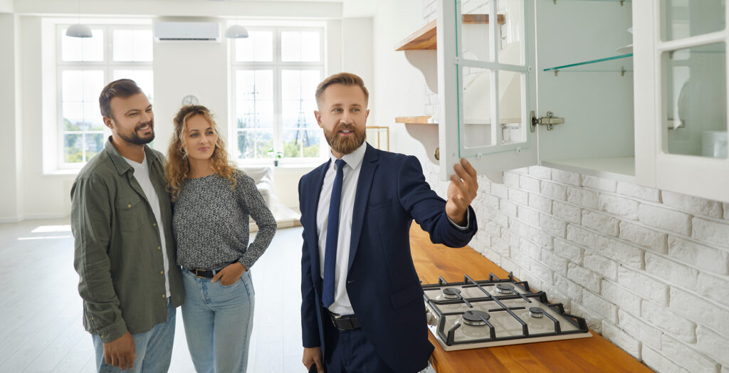 Real estate agent giving first-time homebuyers a tour of their new home with modern wooden kitchen cabinets.