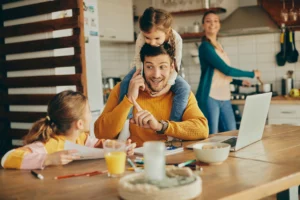 Busy family at home with father working at kitchen table while children interrupt, highlighting challenges of managing insurance and family responsibilities